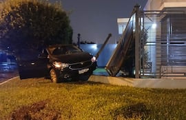 Vehículo negro dañado, portón doblado y césped bien cuidado en un área residencial.