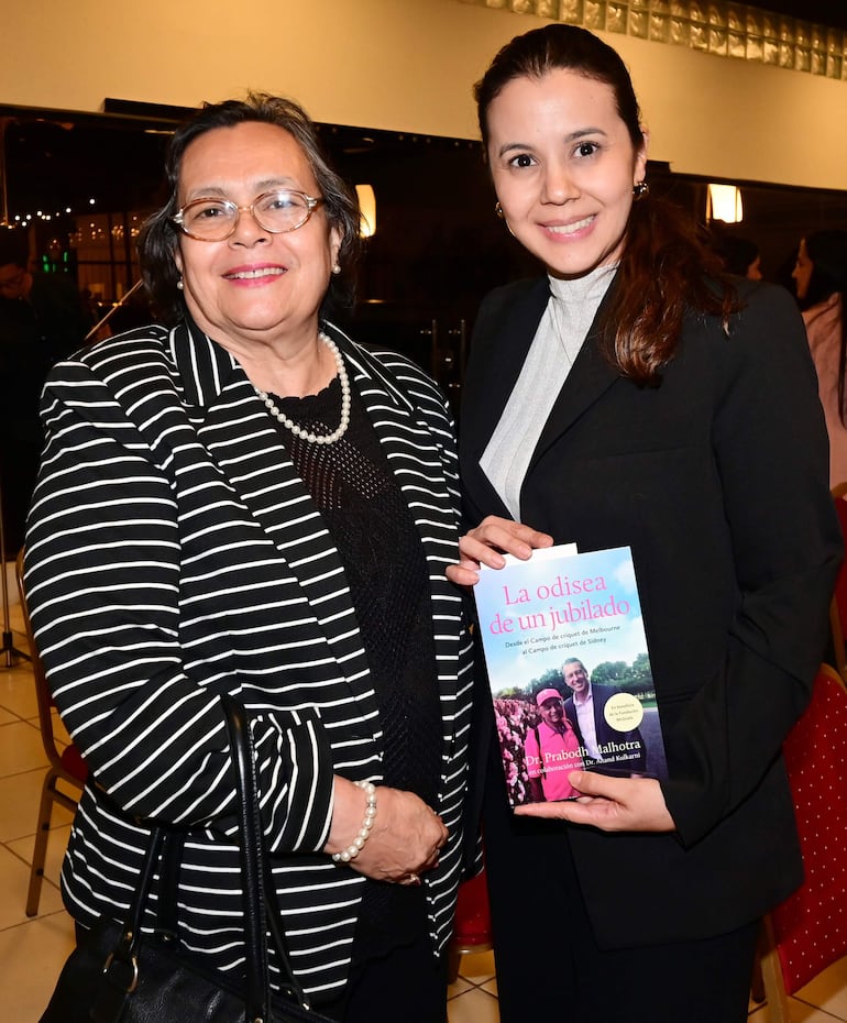 Mujeres sonrientes, una con gafas y blusa negra, otra con blazer negro sosteniendo un libro en un salón iluminado.