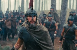 Soldado con casco rojo y capa en primer plano, rodeado de otros guerreros en un bosque nebuloso y tenso.