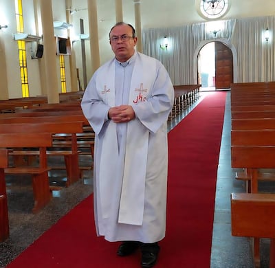 Sacerdote Ismael Obregón Olmedo, vestido con túnica blanca, de pie en el pasillo de una iglesia, con las manos juntas y mirando a la cámara.