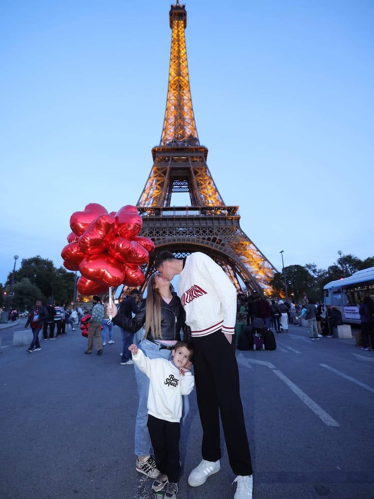 Mujer con globos rojos en forma de corazón, hombre alto inclinado para besarla, niño con sudadera blanca sonriente, Torre Eiffel de fondo.