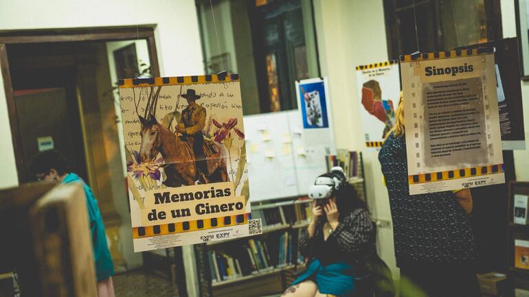 Persona en primer plano usando realidad virtual, con cartel en mano y ambiente cultural decorado con carteles artísticos.