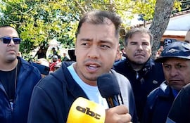 Miguel Prieto Vallejos con micrófono en camiseta blanca y abrigo negro, rodeado de hombres con expresiones serias y atentas.