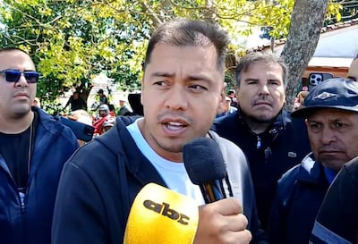 Miguel Prieto Vallejos con micrófono en camiseta blanca y abrigo negro, rodeado de hombres con expresiones serias y atentas.
