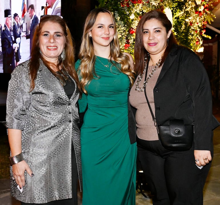 Tres mujeres sonrientes: una en vestido verde, otra en chaqueta plateada y la última en suéter marrón, rodeadas de flores y luces.