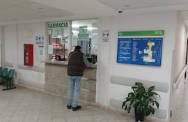 Hombre de espaldas en camisa a cuadros consulta en la farmacia del hospital, con asientos vacíos y carteles de bioseguridad en el entorno.