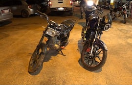 Dos motocicletas apagadas, una negra con naranja y otra negra y azul con rojo, expuestas en un estacionamiento iluminado.