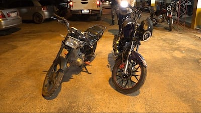 Dos motocicletas apagadas, una negra con naranja y otra negra y azul con rojo, expuestas en un estacionamiento iluminado.