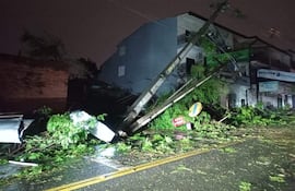 Poste de luz caído y escombros en una calle urbana, mientras un edificio mantiene sus luces encendidas en el fondo.
