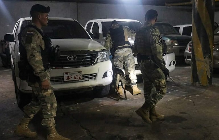 Tres hombres armados en uniforme militar, uno caminando con un arma, otro agachado junto a un vehículo blanco, mientras un perro inspecciona el área.