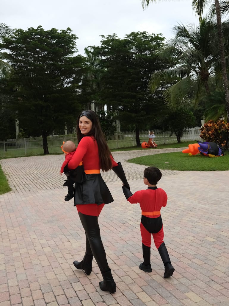 La imagen muestra a una mujer y dos niños en un ambiente exterior, probablemente en un parque. La mujer, que lleva un atuendo rojo y negro, parece estar caminando hacia la izquierda mientras sostiene a un bebé en brazos. El niño, que se encuentra a su lado, también viste un traje rojo similar, haciendo eco de los colores de su acompañante.
Hay elementos decorativos en el fondo, como figuras de Halloween que sugieren un evento festivo o de disfraces. El entorno es verde y arbolado, lo que indica un espacio recreativo. No se observan textos, logos o elementos deportivos, por lo que no se identifican equipos de fútbol ni figuras públicas reconocibles. La acción principal es la caminata de la mujer y los niños, con un ambiente alegre y lúdico.