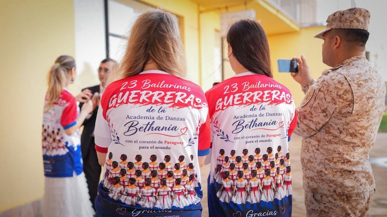 Tres bailarinas de espaldas, vestidas con camisetas que dicen 'GUERRERAS', conversan con un hombre en traje oscuro en un evento al aire libre.