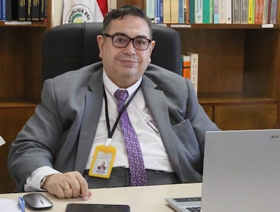 Carlos Mendoza, de traje gris y corbata púrpura, sentado en su escritorio con laptop y teléfono móvil.