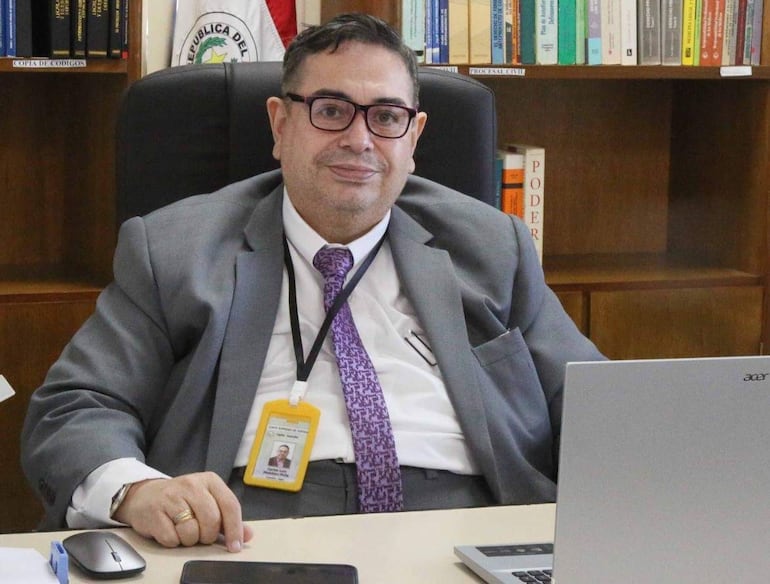 Carlos Mendoza, de traje gris y corbata púrpura, sentado en su escritorio con laptop y teléfono móvil.