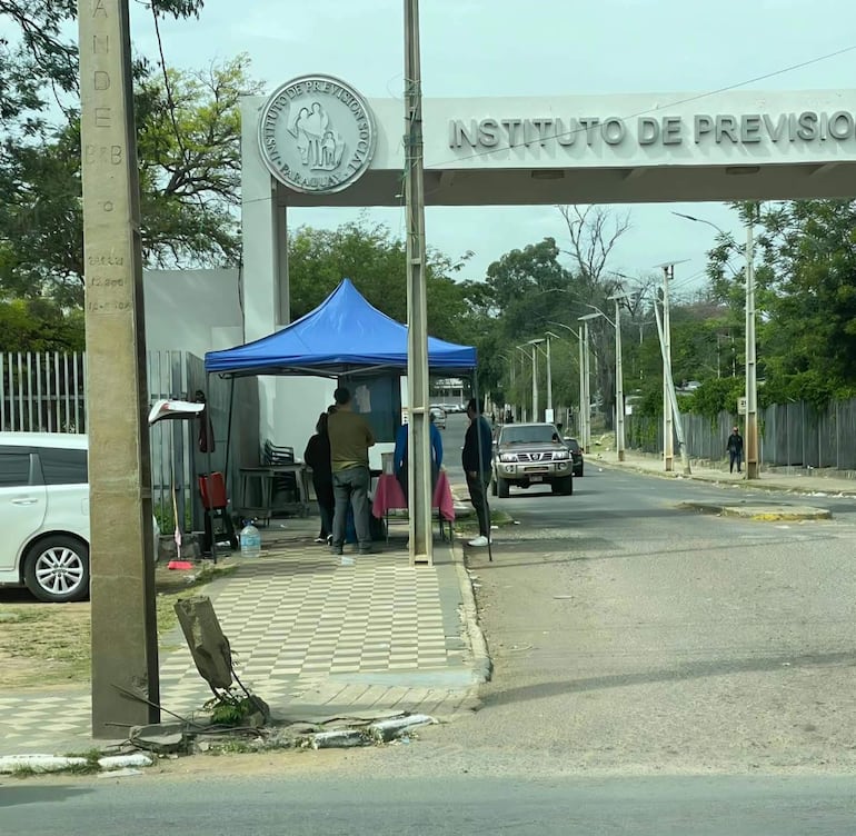 Cinco personas, algunas de pie y otras agachadas, bajo una carpa azul frente al Hospital Central del IPS, con un ambiente tranquilo.