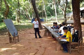 Alumnos de una escuela indígenas dan clases bajo árboles y utilizan un banco rústico como mesa pedagógica.