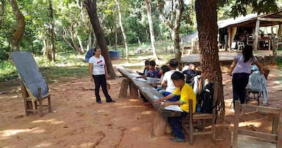 Alumnos de una escuela indígenas dan clases bajo árboles y utilizan un banco rústico como mesa pedagógica.