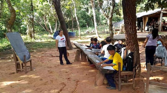 Alumnos de una escuela indígenas dan clases bajo árboles y utilizan un banco rústico como mesa pedagógica.