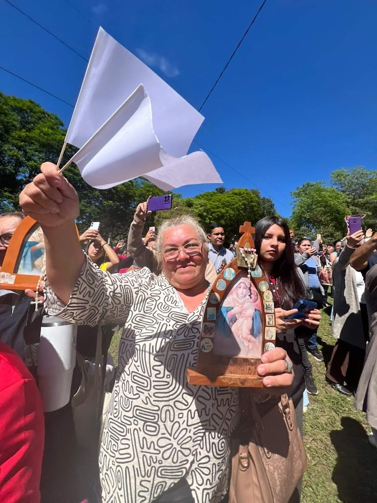 Mujer rubia con bandera blanca y imagen religiosa en festividad, rodeada de público con teléfonos en un ambiente festivo.