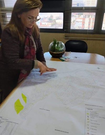 Rosa Vacchetta, mujer con gafas y prenda oscura, sostiene un mapa en la mesa rodeada de documentos y plantas en oficina.