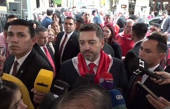 Pedro Alliana en el centro, con expresión seria, rodeado de comunicadores en trajes oscuros y bufandas rojas.