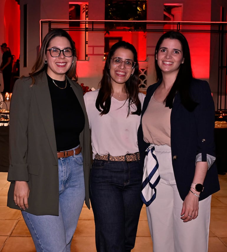 Tres mujeres sonrientes en evento, una con blusa negra y chaqueta verde, otra en blusa clara con cinturón animal y la tercera con blusa beige y bufanda.