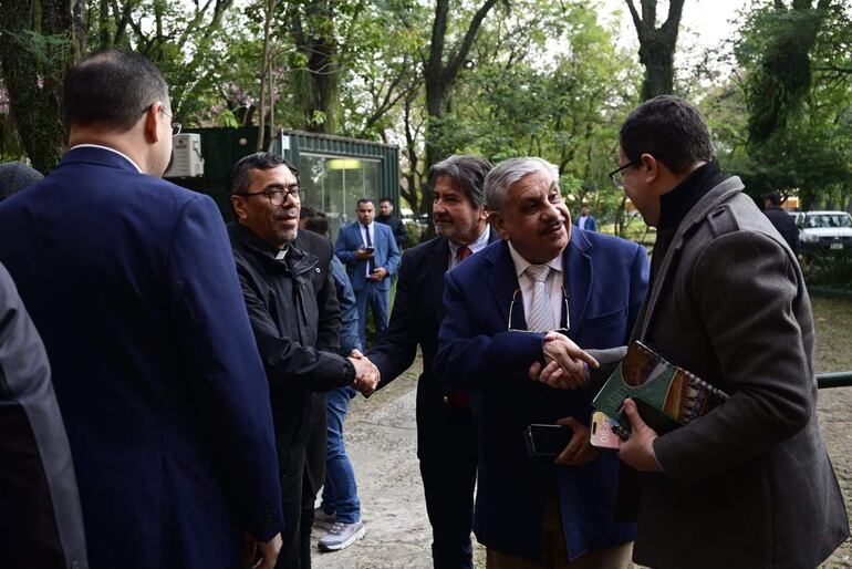 Dos hombres en primer plano se saludan; uno lleva abrigo oscuro y gafas, el otro traje claro con gafas de sol sobre la cabeza.