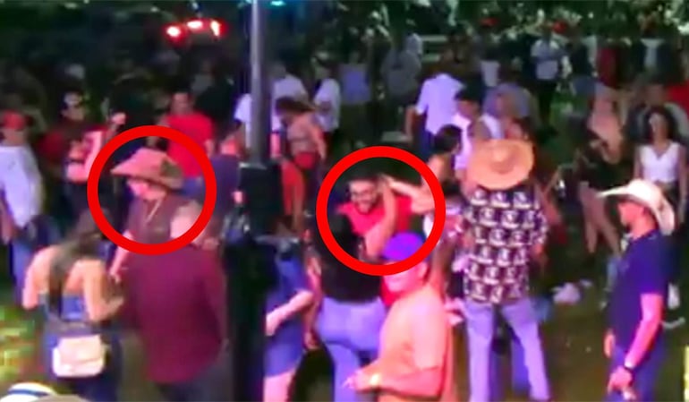 Hombre con sombrero grande y otro con gafas y camiseta roja, sonriendo en un ambiente animado de celebración.
