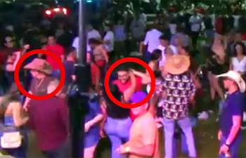 Hombre con sombrero grande y otro con gafas y camiseta roja, sonriendo en un ambiente animado de celebración.