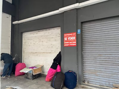 Dos figuras en chaquetas oscuras, una de espaldas, junto a un cartel rojo que dice 'SE VENDE' en un entorno urbano desolado.