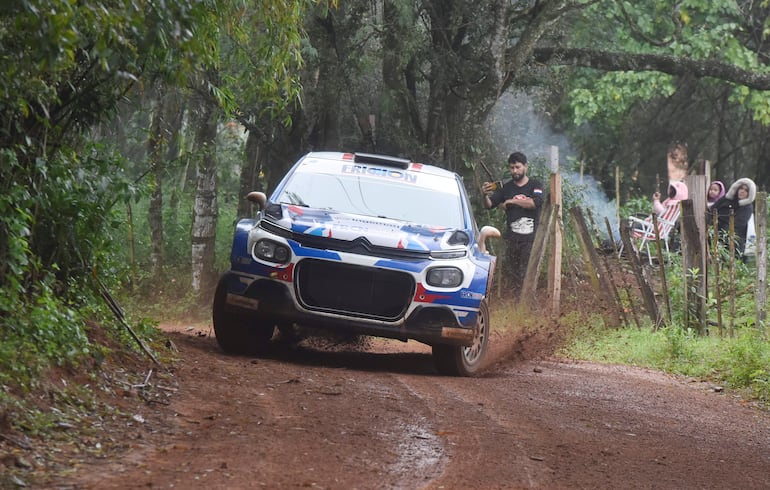 Coche de rally azul en curva, rodeado de vegetación, con persona al fondo grabando la competencia.