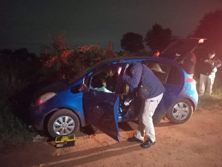 Tres policías en uniforme observan un coche azul con la puerta abierta, revisando el interior en una escena oscura y poco iluminada.
