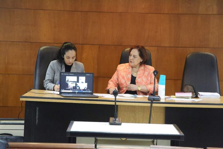 Dos mujeres sentadas en una audiencia, una con camisa gris y auriculares, la otra en blazer naranja, concentradas en documentos.