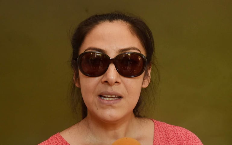 Mujer con gafas oscuras y blusa rosada, hablando a la cámara en un ambiente interior verde.