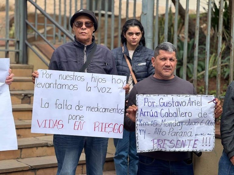 Tres personas en protesta con carteles sobre la falta de medicamentos, dos sosteniendo reclamos, una con expresión de atención. Ambiente exterior.