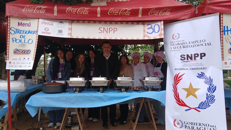Grupo de personas en un stand de comida, incluyendo a un hombre en traje oscuro y mujeres con cofia, en ambiente festivo.