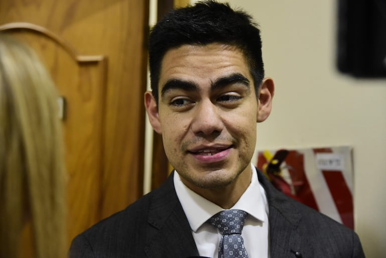 Hombre con cabello corto y oscuro en traje oscuro y corbata, hablando en un entorno formal con muebles de madera al fondo.
