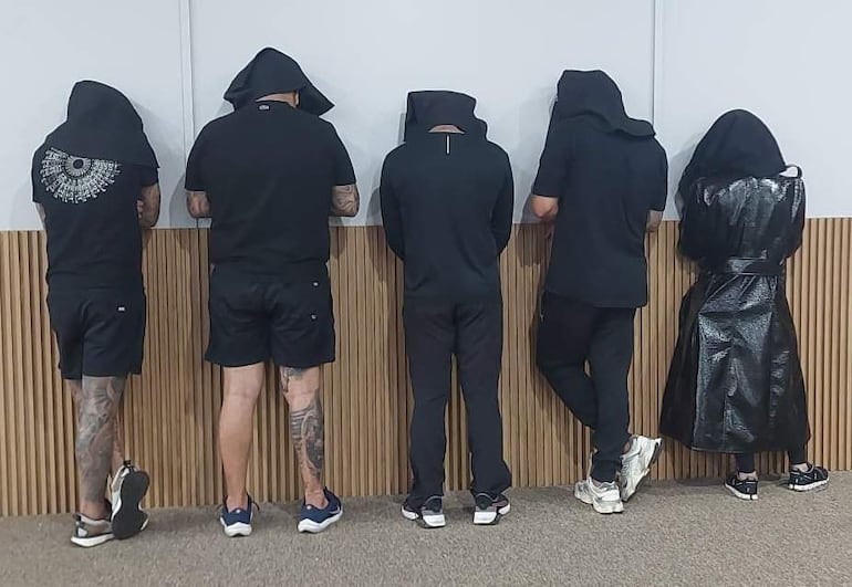 Cinco personas de espaldas en prendas oscuras, esperando en un ambiente interior con pared de madera y alfombra.