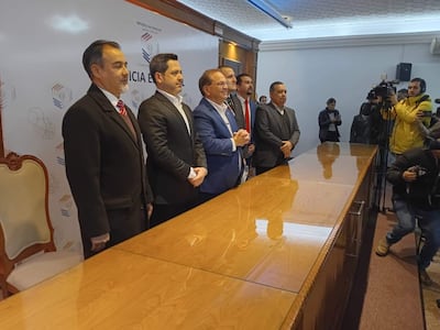 Camilo Pérez en traje oscuro frente a mesa, rodeado de hombres con vestimenta formal, en un ambiente de conferencia.