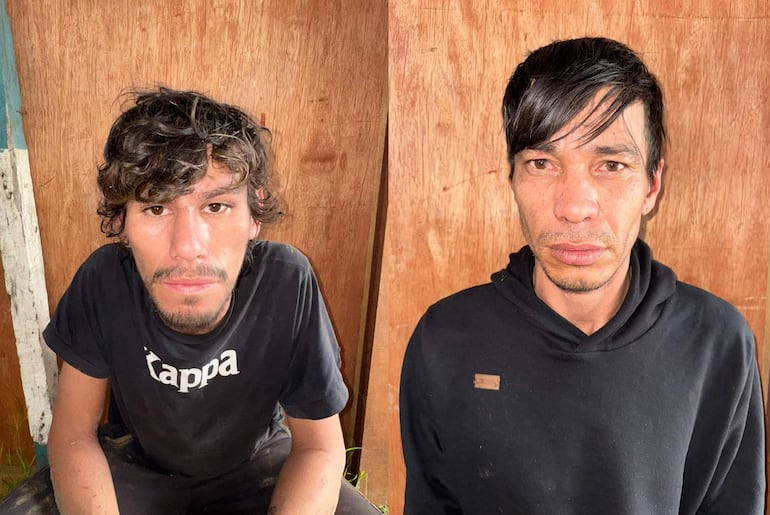 Dos hombres, uno con camiseta negra que dice 'Kappa' y otro en sudadera negra, miran seriamente a la cámara en un ambiente interior.
