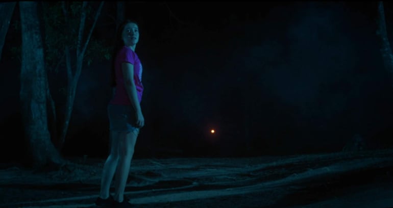 Joven con camiseta rosa y pantalones cortos grises, mirando con inquietud hacia una luz lejana en un bosque oscuro.