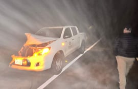 Hombre de espaldas con chaqueta oscura y pantalones claros, caminando lejos de la camioneta blanca dañada en la carretera nocturna.