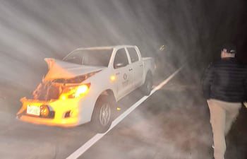 Hombre de espaldas con chaqueta oscura y pantalones claros, caminando lejos de la camioneta blanca dañada en la carretera nocturna.