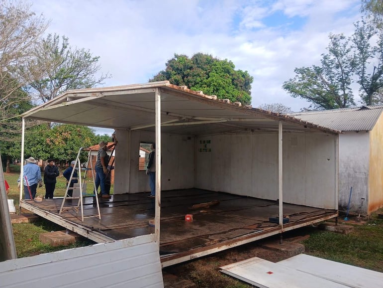 Seis personas, algunas con guantes y sombreros, trabajan en una construcción deteriorada rodeada de árboles y cielo nublado.