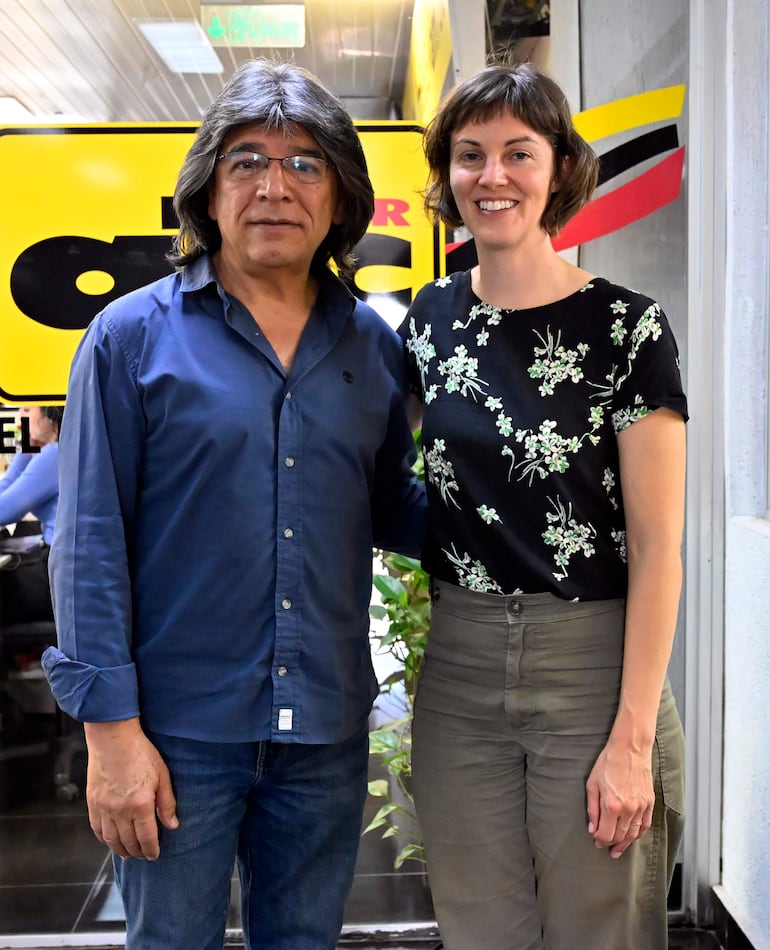 Mujer con blusa negra con flores y pantalones claros, junto a hombre con camisa azul y jeans, ambos sonriendo en un ambiente profesional.