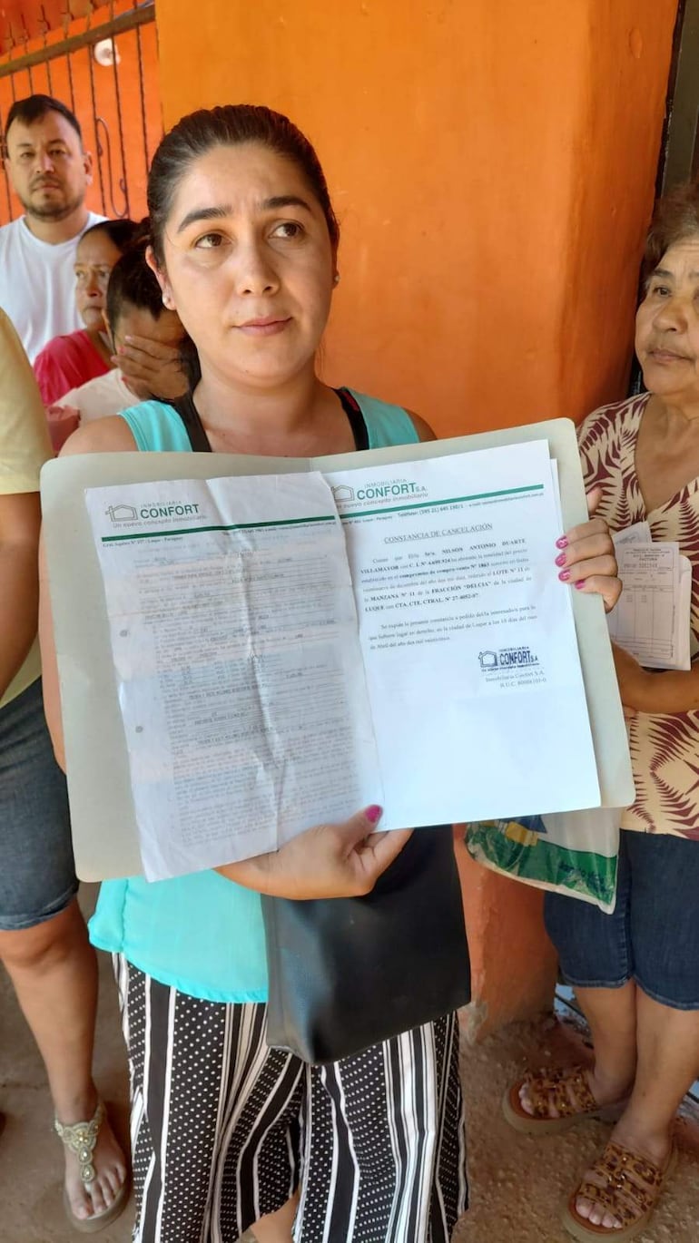 Mujer joven con blusa celeste y falda rayada sostiene documento, en medio de otros cuatro vecinos con expresiones serias.