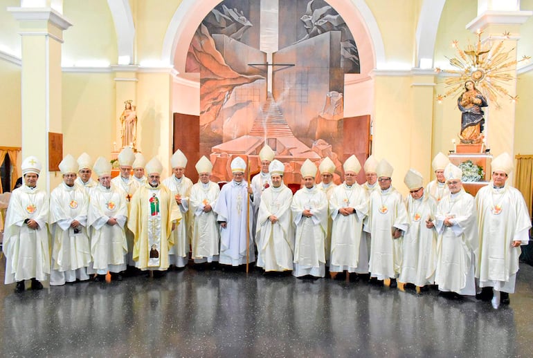 Clérigos vestidos con túnicas blancas y mitras, alineados ante un mural con una cruz, en un ambiente solemne.