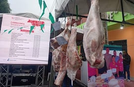 Puesto de carne en un entorno urbano, colgado en un toldo con precios visibles. Un grupo de personas asiste al fondo.