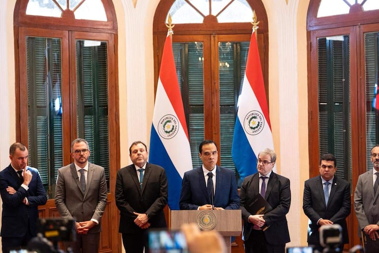Seis hombres en trajes formales en un ambiente elegante, con la bandera de Paraguay de fondo, durante un anuncio importante.