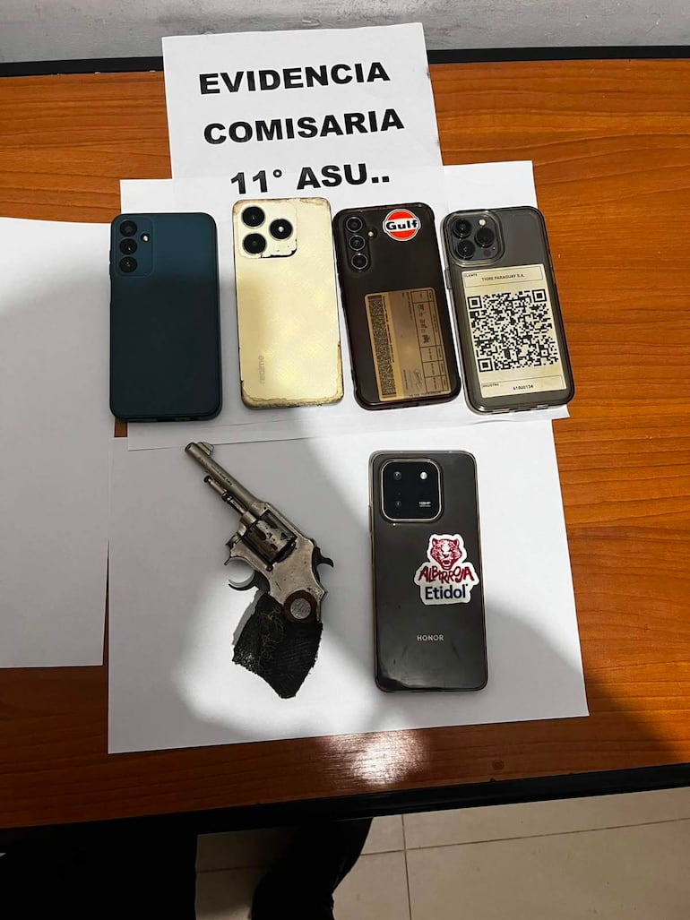 Mesa con cinco teléfonos móviles y un revólver, en fondo blanco, marcados como evidencia policial en comisaría.